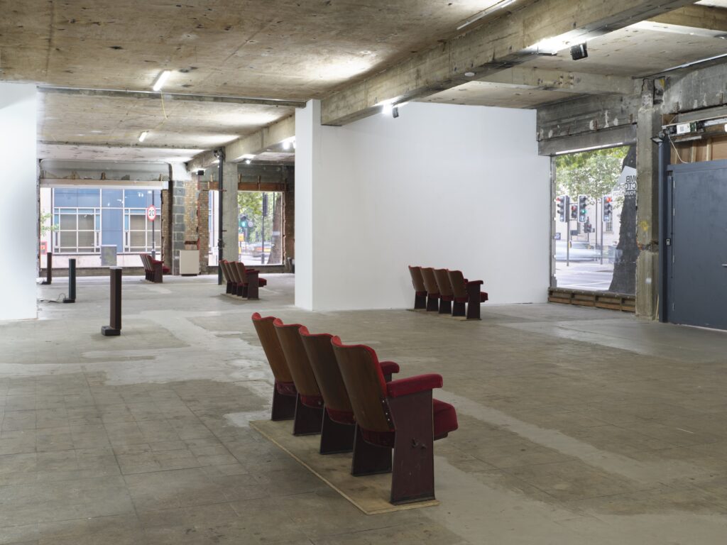 An industrial-style gallery space with concrete floors and ceiling features several rows of red theater seats arranged sparsely. Large windows and doors provide natural light and views of the street outside.