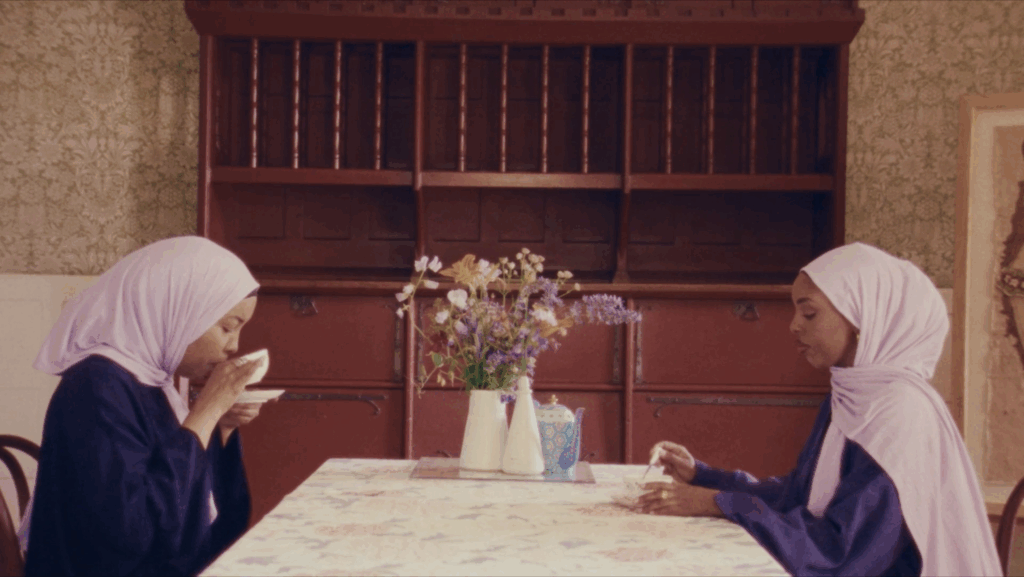 Two women wearing light pink headscarves sit across from each other at a floral-covered table, one drinking from a cup and the other looking down. A vase of flowers is in the center; wooden shelves are in the background.