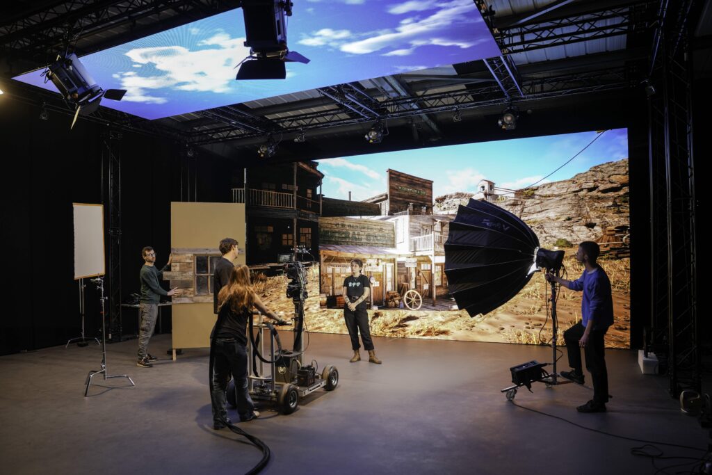 Four people operate cameras and lights on a film set with a large digital screen displaying an Old West town and desert scene in the background. The studio is filled with lighting and filming equipment.