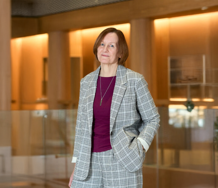 A woman wearing a gray plaid suit and maroon top stands indoors in a modern, warmly lit building with wooden walls and glass railings.