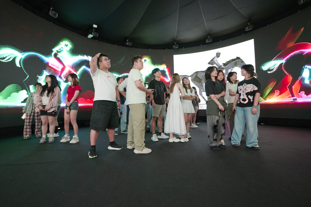A group of people stands inside a dark room with colorful, glowing digital art of mythical creatures displayed on a large curved screen behind them. Most are casually dressed and engaged in conversation or looking around.
