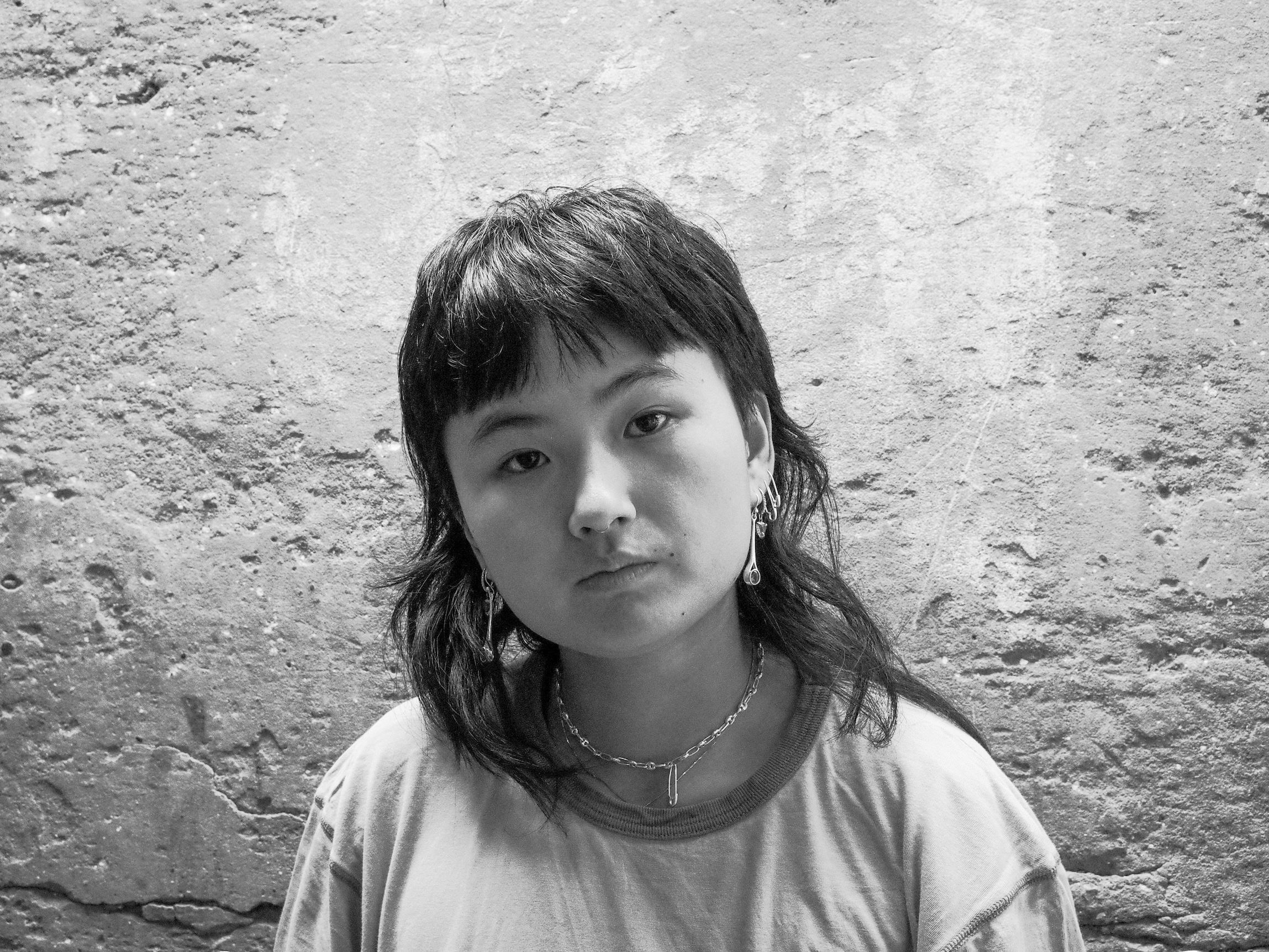 A person with a mullet hairstyle, wearing earrings and a necklace, looks directly at the camera with a neutral expression. The black-and-white image evokes an east gallery vibe against a textured, rough concrete wall.