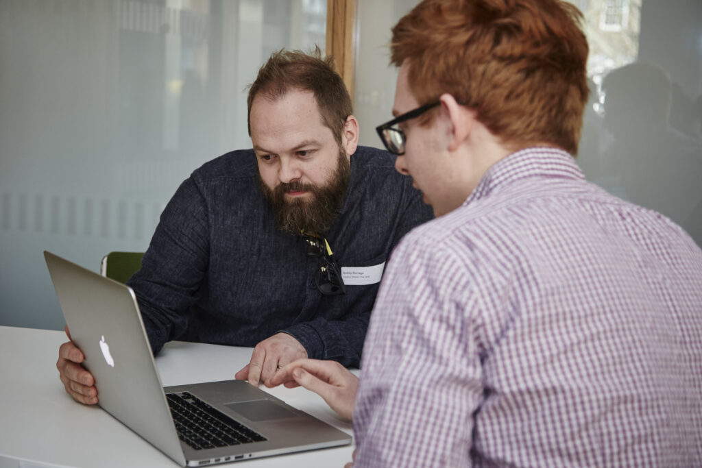 Two men sit at a table, closely looking at a laptop screen. One man is pointing at the laptop while the other attentively follows along. Both appear to be engaged in discussion in a bright office setting.