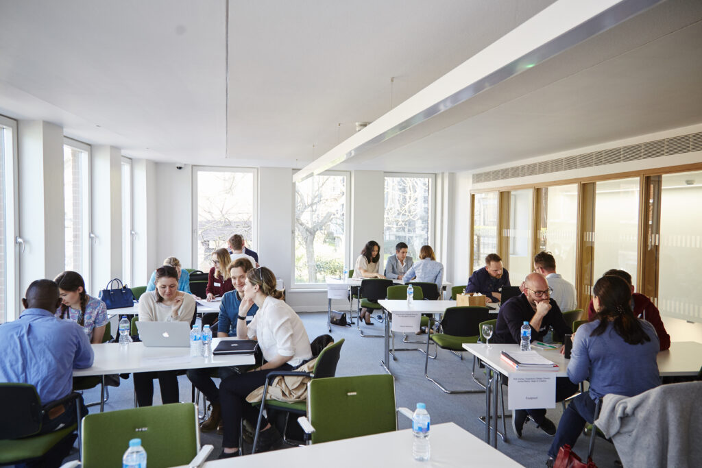 People sit in small groups at tables in a bright, modern meeting room with large windows, talking and working on laptops and documents. Bottles of water and folders are on the tables.