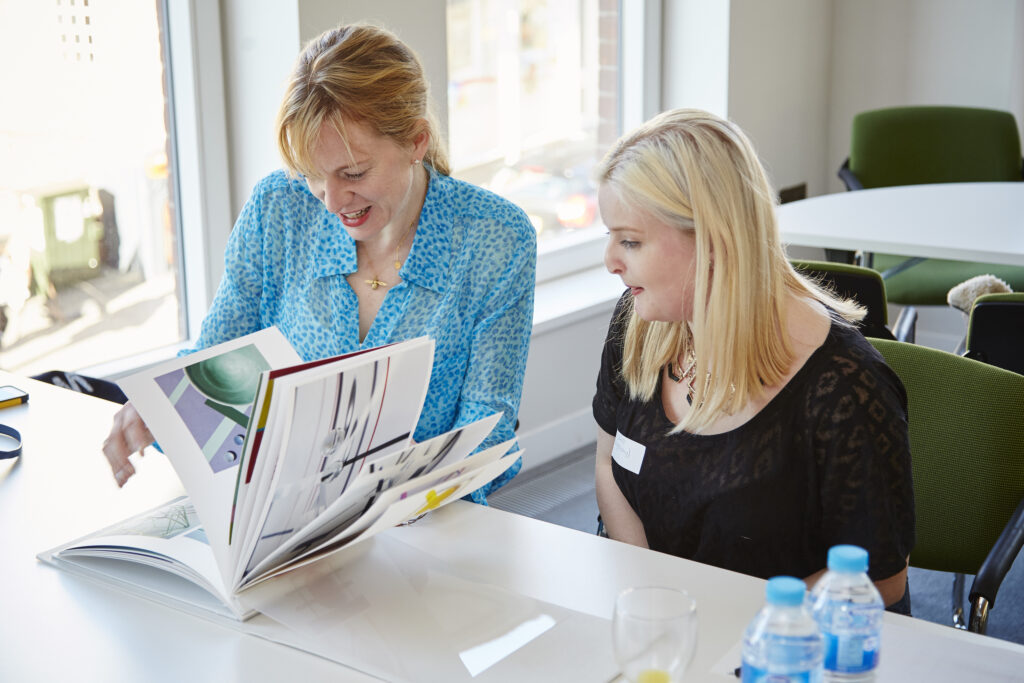 Two women sit at a table looking at an open book together. One woman is wearing a blue shirt, the other a black shirt. Bottled water and empty glasses are on the table in a bright room with large windows.