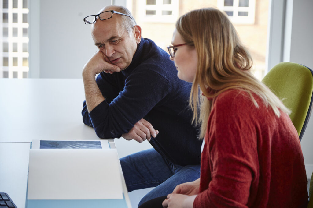 An older man and a younger woman sit at a table in a bright office, looking at documents together. The man has glasses on his head and appears to be listening intently to the woman, who is talking.