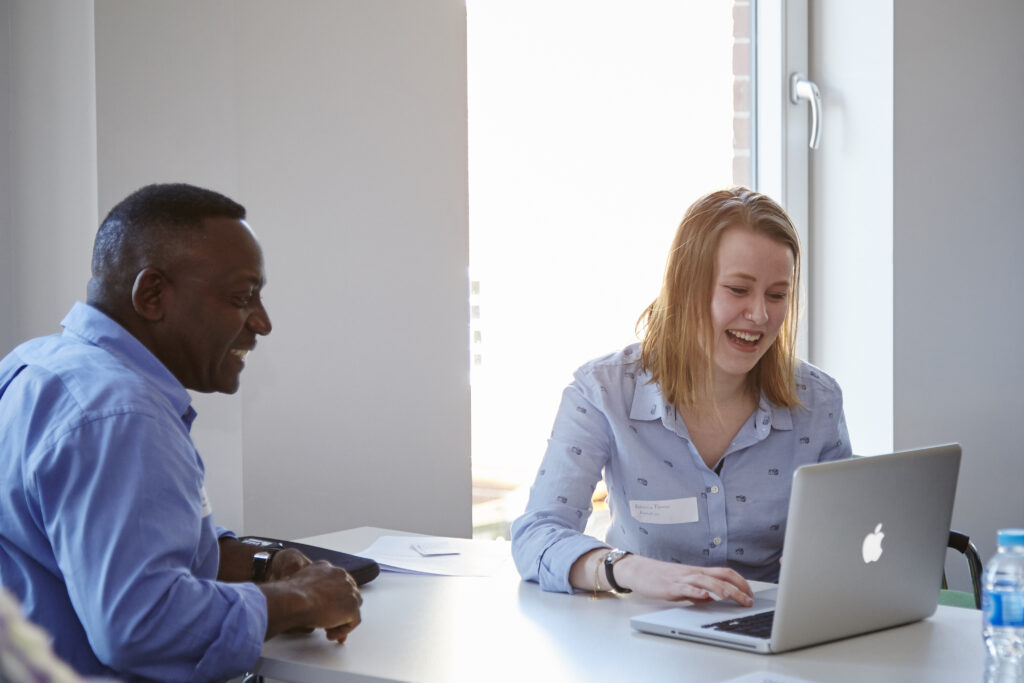 Two people, one man and one woman, sit at a table in a bright room. The woman is using a laptop and smiling, while the man beside her also smiles. Papers and a water bottle are on the table.
