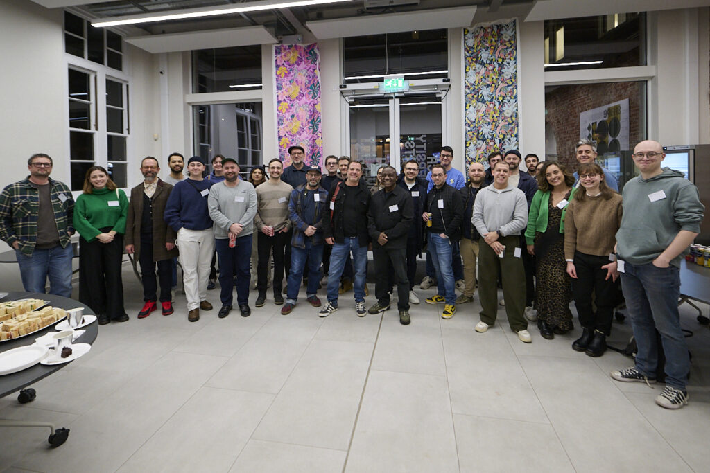 A group of about 25 people stands smiling in a brightly lit room with tall windows, colourful curtains, and refreshments on a table in the foreground. Everyone appears gathered for a group photo at an indoor event.