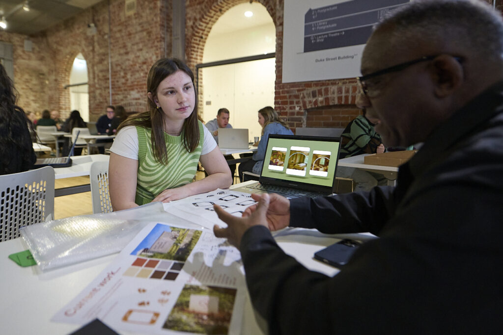 A woman and a man sit across from each other at a table in a modern, brick-walled room, discussing design materials and images, with papers and a laptop in front of them.