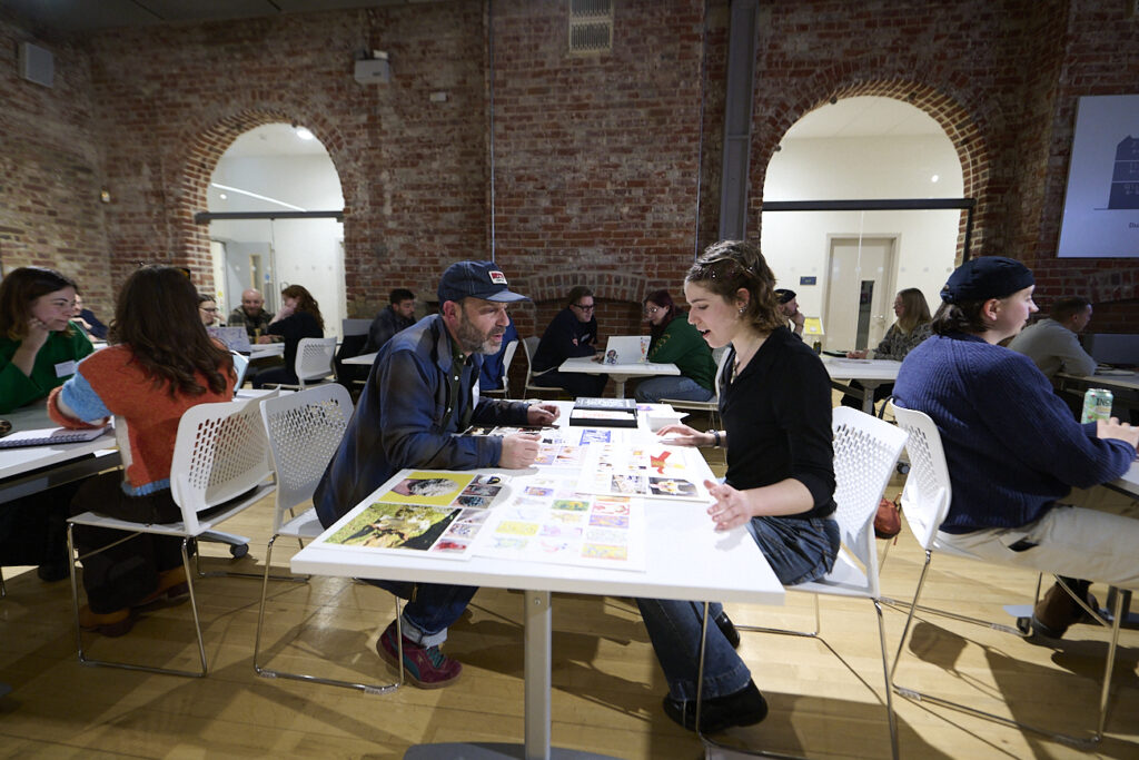 People sit in pairs at tables, some discussing artwork or papers spread in front of them, in a spacious room with exposed brick walls and arched doorways. The atmosphere appears collaborative and creative.