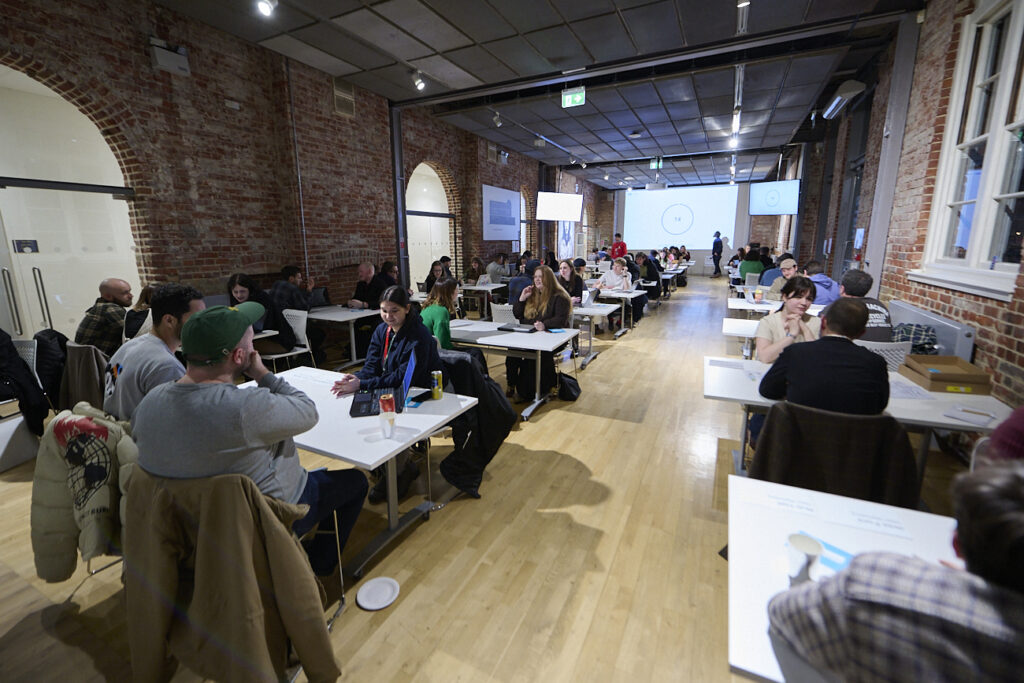 A large room with brick walls and wooden floors, filled with groups of people sitting at tables, talking and working together. A projector screen is visible at the front of the room.