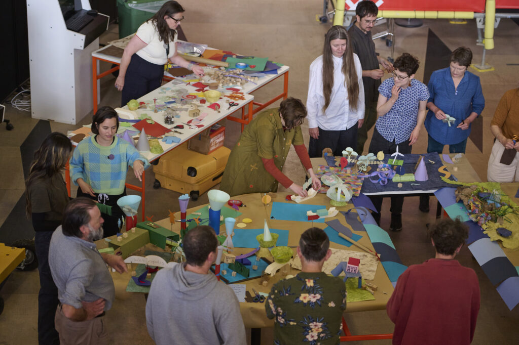 Tales from Imagined Coastal Futures, a workshop as part of the Sainsbury Centre & Norwich University of the Arts' symposium 'Shared Seas: Coastal Encounters'. A group of people stands around tables covered with colourful paper, models, and art supplies, collaboratively working on creative projects in a bright, indoor space.