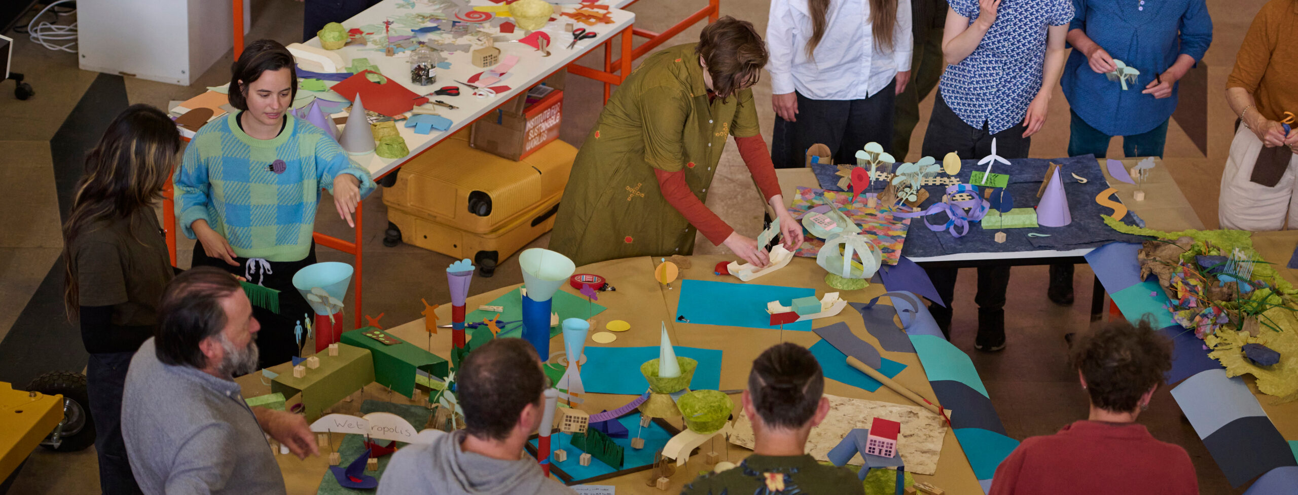 A group of people gather around tables covered with colorful craft materials and paper sculptures, working together on creative art projects in a bright indoor space.