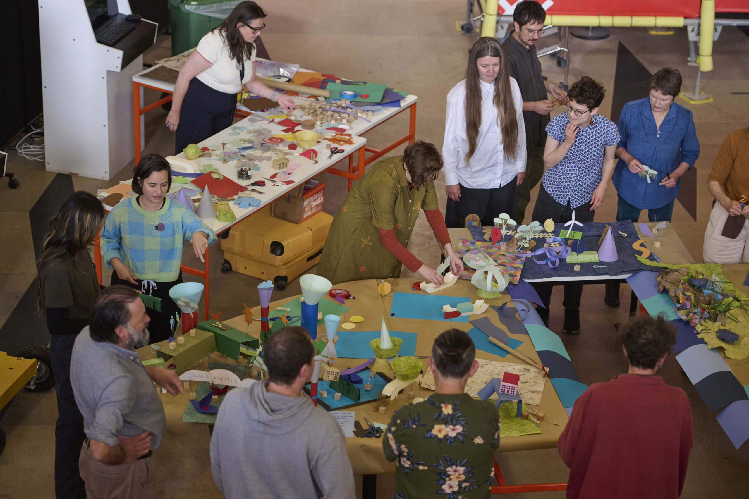 Tales from Imagined Coastal Futures, a workshop as part of the Sainsbury Centre & Norwich University of the Arts' symposium 'Shared Seas: Coastal Encounters'. A group of people stands around tables covered with colourful paper, models, and art supplies, collaboratively working on creative projects in a bright, indoor space.