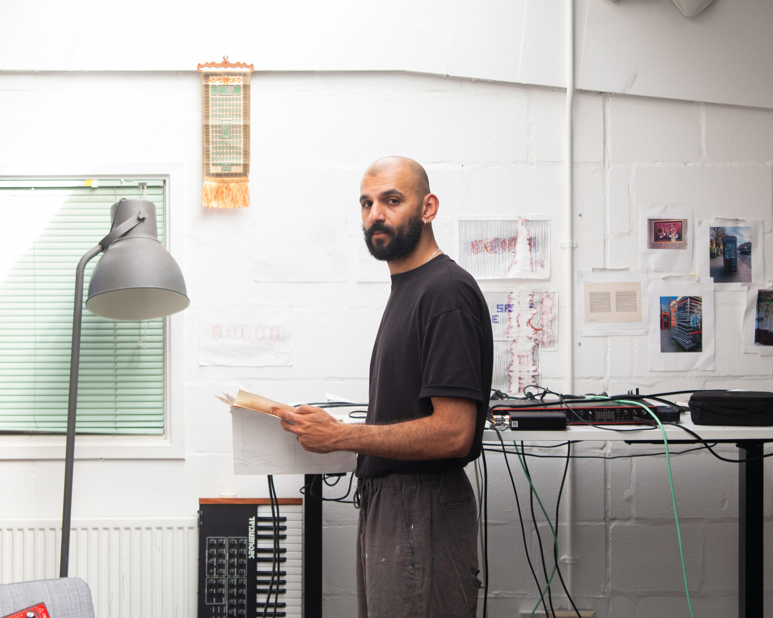 A person with a beard stands indoors holding an open book, looking at the camera. Behind them are electronic equipment, a lamp, a window with blinds, and papers on a white brick wall.