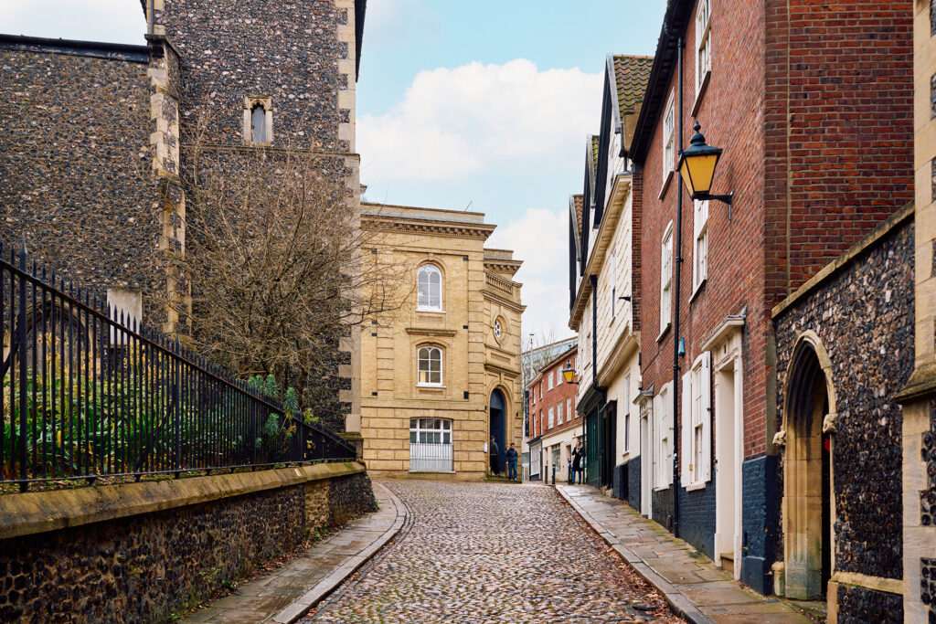 A narrow cobblestone street lined with historic brick and stone buildings curves uphill, with a tall church tower on the left and a yellow lamp hanging from a building on the right under a partly cloudy sky.