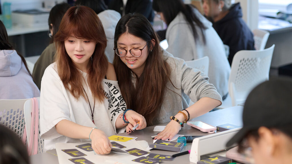 Two young women sit at a table, smiling and working together on an activity with cards. Other people are seated and engaged in similar activities in the background. The setting appears to be a classroom or workshop.