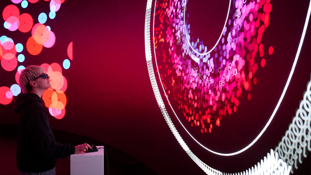 A person with short light hair and sunglasses stands at a console, gazing at a large, brightly lit digital display of concentric circles and multicolored dots against a dark pink and red background.