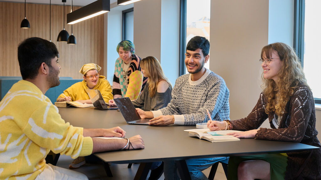 Six young adults sit around a table in a modern, well-lit room, smiling and talking. Some have laptops or notebooks open, suggesting a collaborative study or meeting environment.