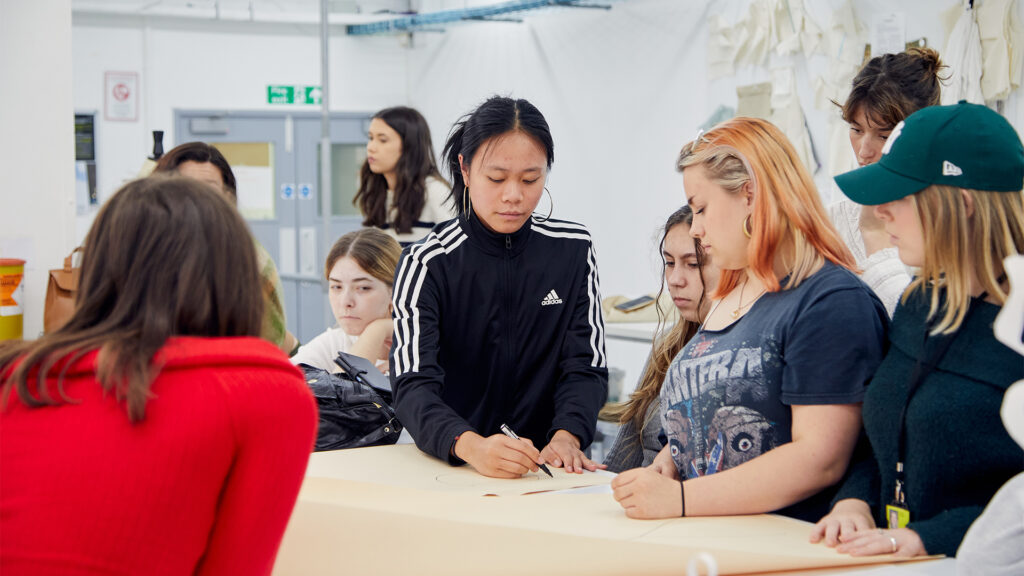 A group of students gathers around a table. One person in a black Adidas jacket draws on a large sheet of paper while others watch attentively in a bright classroom with white walls and pattern pieces hanging in the background.