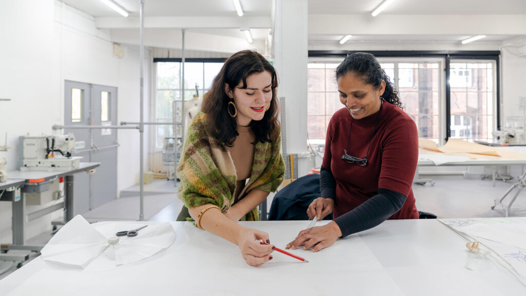 Two women smiling and collaborating at a large white table in a bright, modern studio, reviewing a drawing with pencils and sewing materials visible nearby. Sewing machines are in the background.