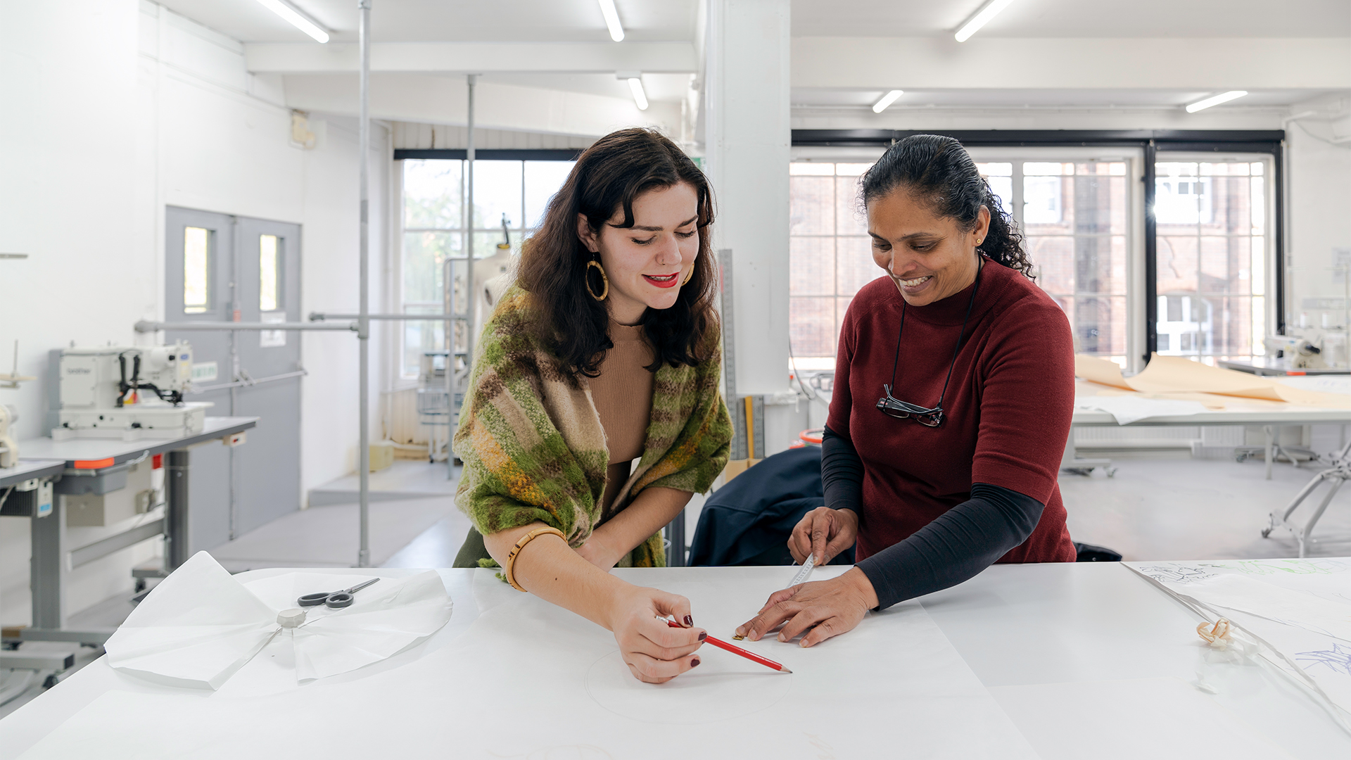 Two women smiling and collaborating at a large white table in a bright, modern studio, reviewing a drawing with pencils and sewing materials visible nearby. Sewing machines are in the background.