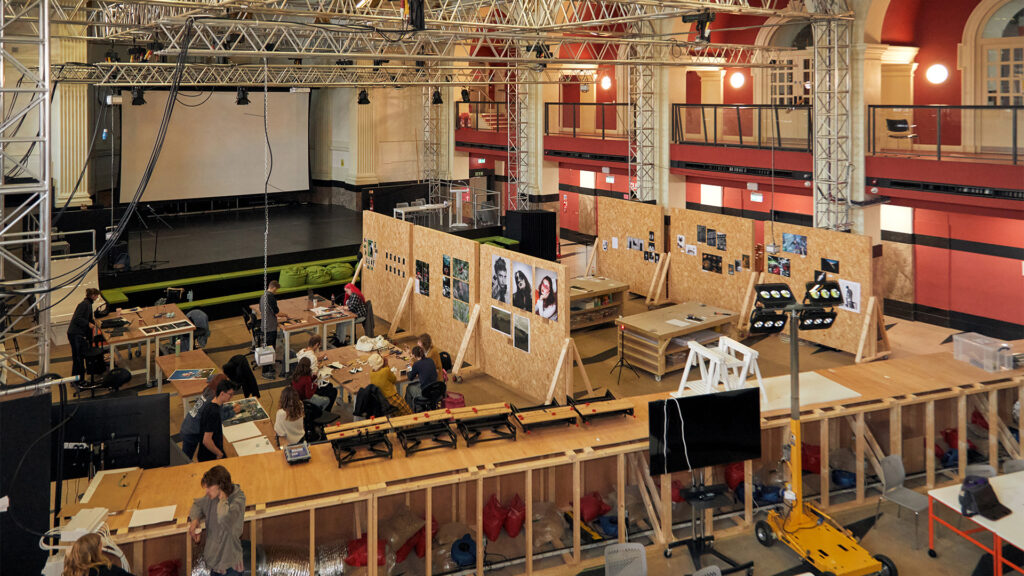 A large, open indoor workspace with people working at tables, surrounded by wooden partitions displaying photos and art. The area is industrial and modern, with exposed beams, stage lighting, and high ceilings.