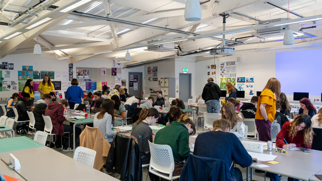 A busy classroom with students sitting at tables, working on creative projects. Several instructors stand and interact with students. The room has bright lighting, white walls, and posters displayed throughout.