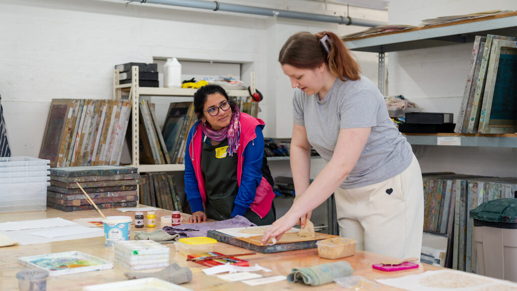 Two women work together in an art studio. One woman stands pressing a print, while the other watches with interest. Art supplies, paint, and papers are spread across a large table; shelves with materials are in the background.