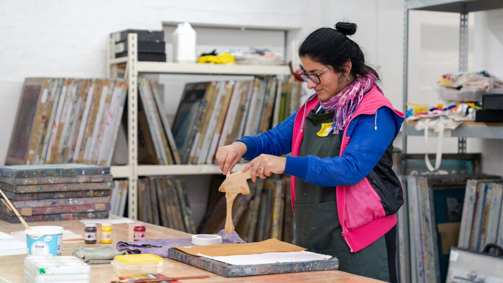 A woman wearing glasses, a pink jacket, and an apron works on an art project in a studio, handling brown material over a white canvas with paints and art supplies on the table around her. Shelves and art frames are in the background.