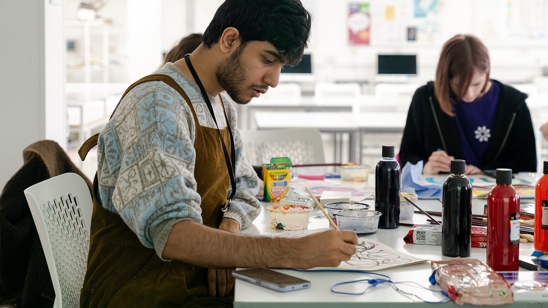 A person wearing a brown apron sits at a table, drawing or painting on paper with art supplies and paint bottles around them. Another person works in the background in a bright, modern room.