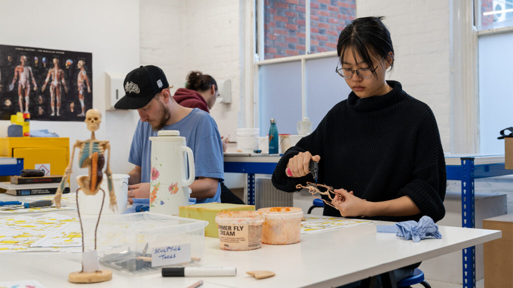 Two people work at a table covered with art supplies and anatomical models. The person in the foreground shapes a small sculpture, while another works in the background. The setting appears to be a creative studio or classroom.