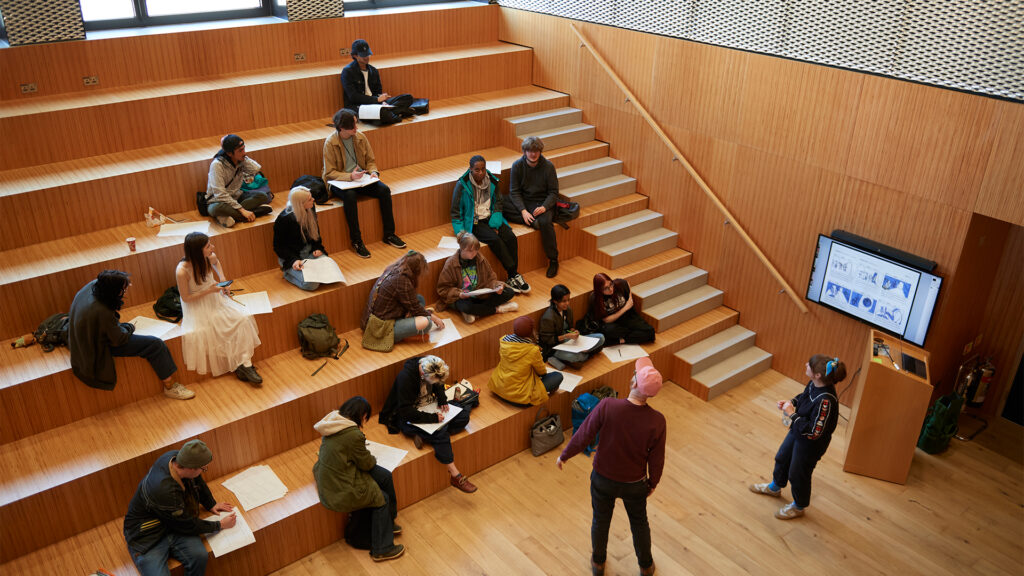 A group of students sit on wooden tiered seating in a modern classroom, listening and taking notes while two people stand near a screen displaying images and diagrams.