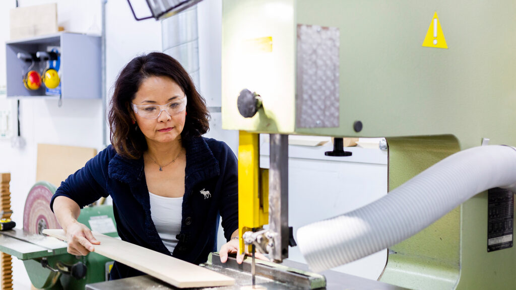 A woman wearing safety glasses operates a bandsaw, cutting a piece of wood in a well-lit workshop with tools and equipment in the background.