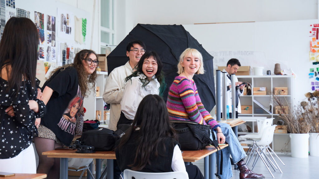 A group of young adults laugh and chat together in a bright art studio, surrounded by creative materials, posters, and equipment. Some are seated at tables, while others stand, all appearing joyful and engaged.