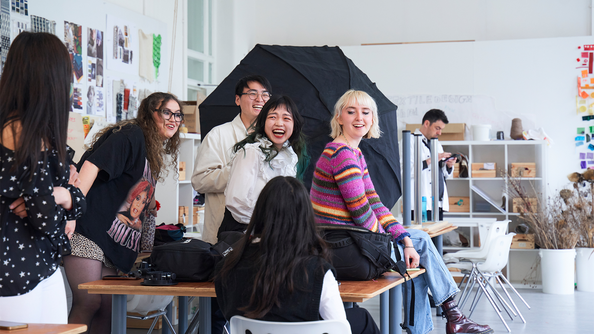 A group of young adults laugh and chat together in a bright art studio, surrounded by creative materials, posters, and equipment. Some are seated at tables, while others stand, all appearing joyful and engaged.