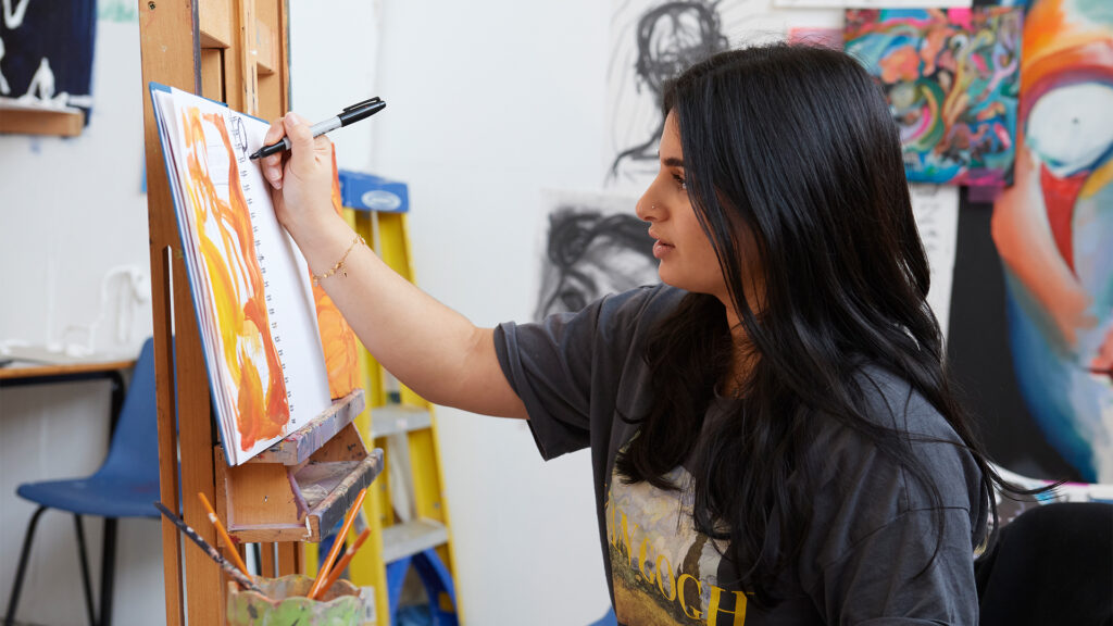 A woman with long dark hair is painting on a canvas at an easel in an art studio, surrounded by colorful artwork and art supplies.