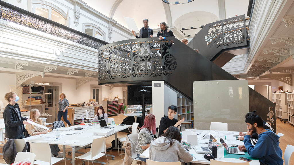 A group of students work at tables with laptops and papers in a spacious, modern classroom with ornate railings and a central staircase. Two people stand talking on the stairs above the students.