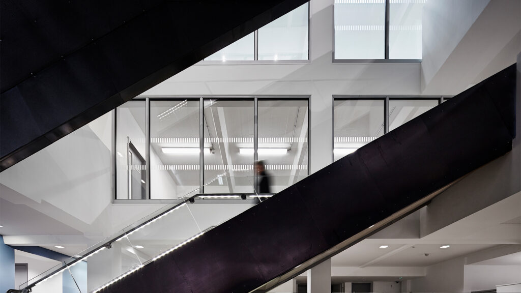 Modern interior with large black diagonal escalators, glass walls, and bright ceiling lights. A blurred person is visible behind the glass, creating a sense of motion in a sleek, geometric architectural space.