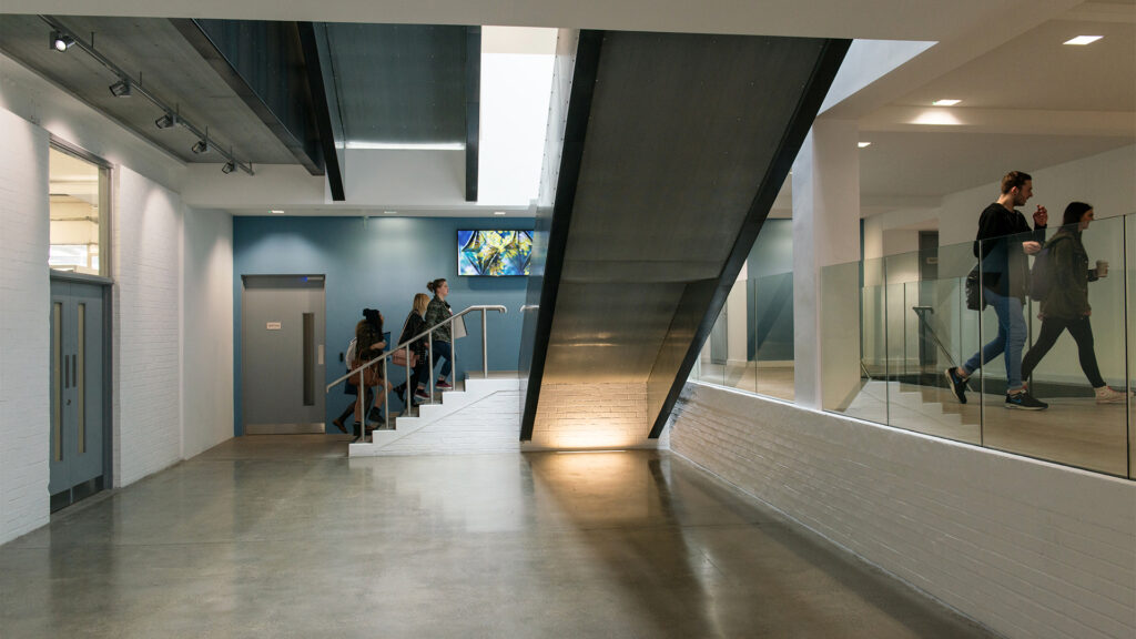 A modern building interior with people walking and a group ascending stairs under a dark metal staircase; the space features glass railings, white brick walls, and polished concrete floors.
