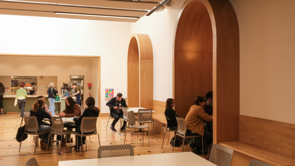 A modern cafeteria with groups of people sitting at tables and in wooden alcove booths along the wall, while others stand near the counter in the background. The space is bright with light wood and white accents.