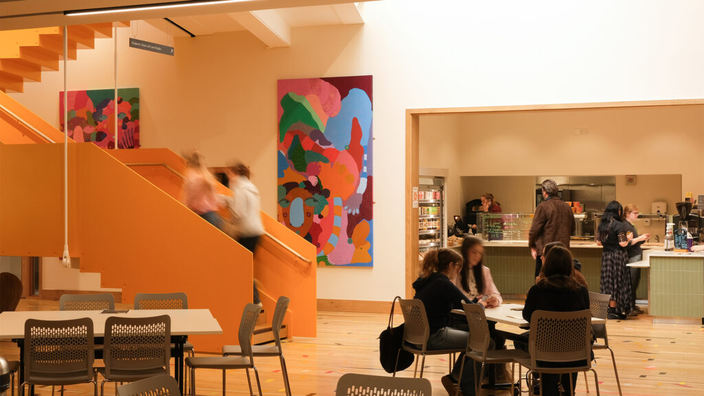 A brightly lit café with people sitting at tables, others ordering at a counter, and a few walking up an orange staircase. Colorful abstract artwork hangs on the walls.