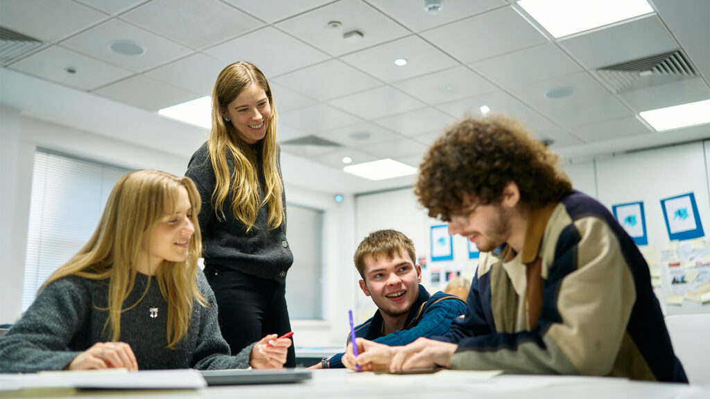 Four young adults work together at a table in a bright, modern classroom. Three are seated and writing or talking, while one stands and smiles, engaging with the group. Papers and pens are on the table.