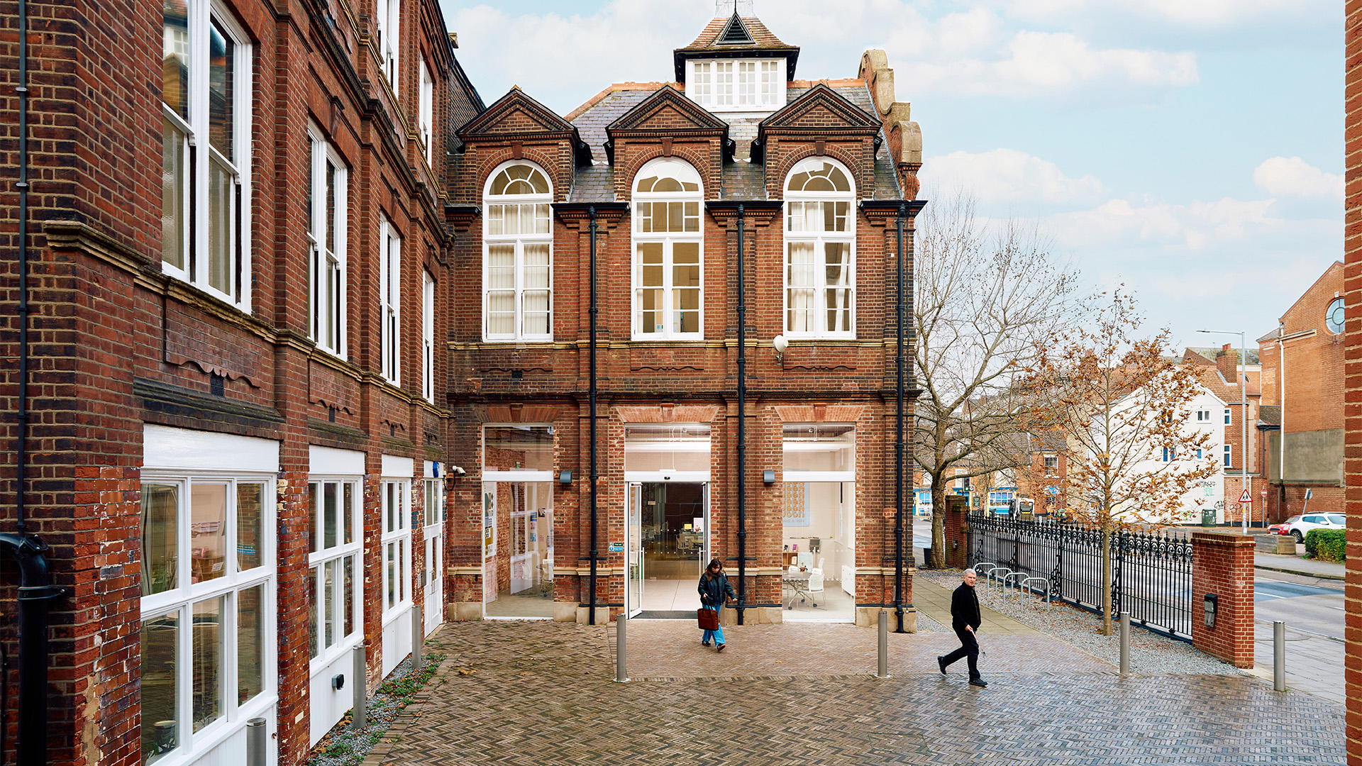 A red-brick building with large arched windows and a modern glass entryway, people walking on a cobblestone path, and trees and houses visible in the background.