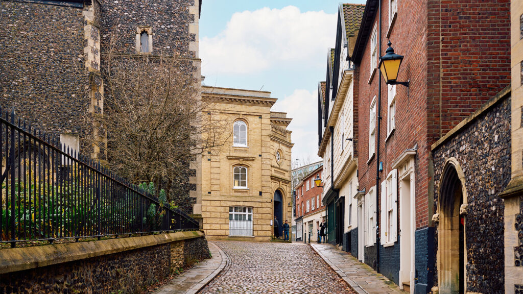 A cobblestone street lined with historic brick and stone buildings curves uphill, with a wrought-iron fence and greenery on the left and a vintage-style streetlamp on the right under a partly cloudy sky.