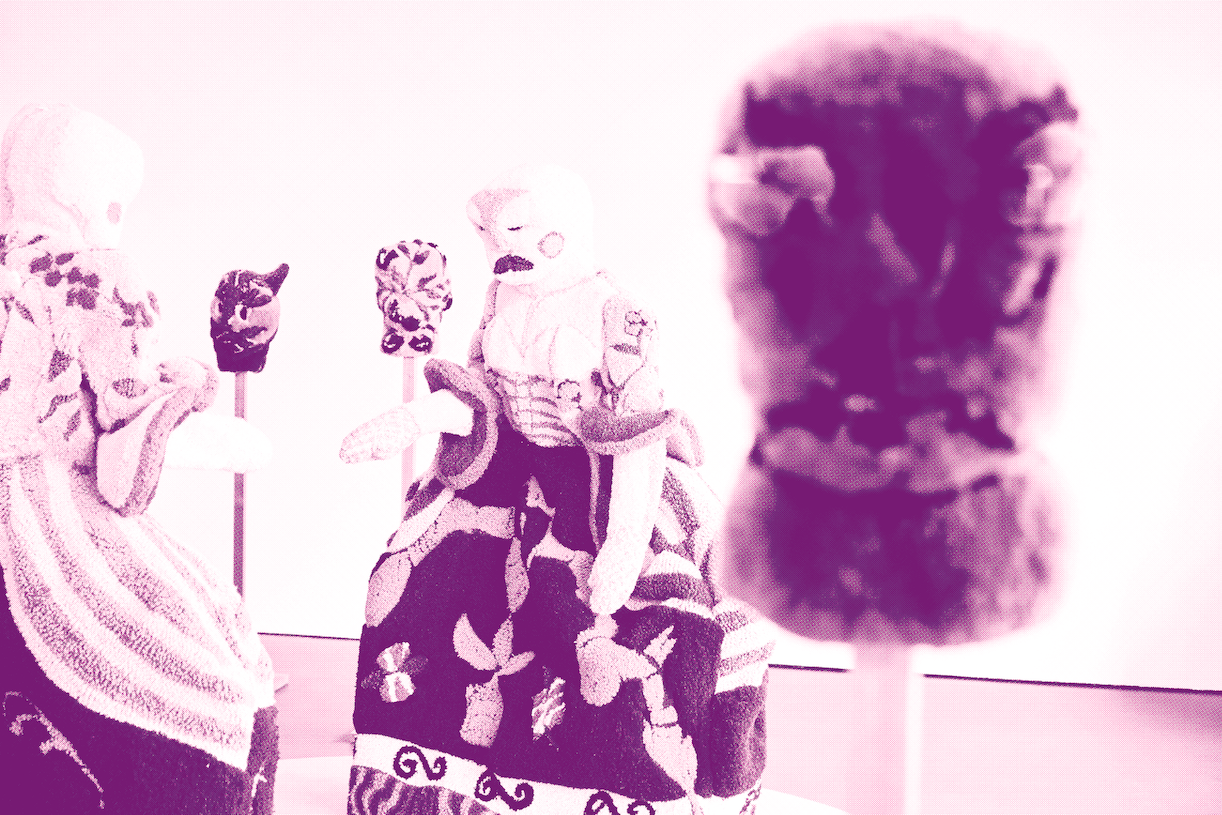 Three fabric puppets with detailed clothing and patterns are positioned in conversation, while a fourth puppet appears blurred in the foreground. The image is tinted with a soft pink hue.