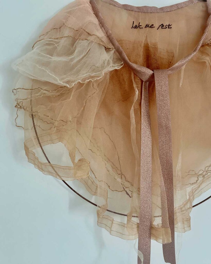 A delicate, sheer, peach-colored ruffled collar with embroidered text 'let me rest' on the neckline and pink ribbon ties, hanging on a thin circular wire.