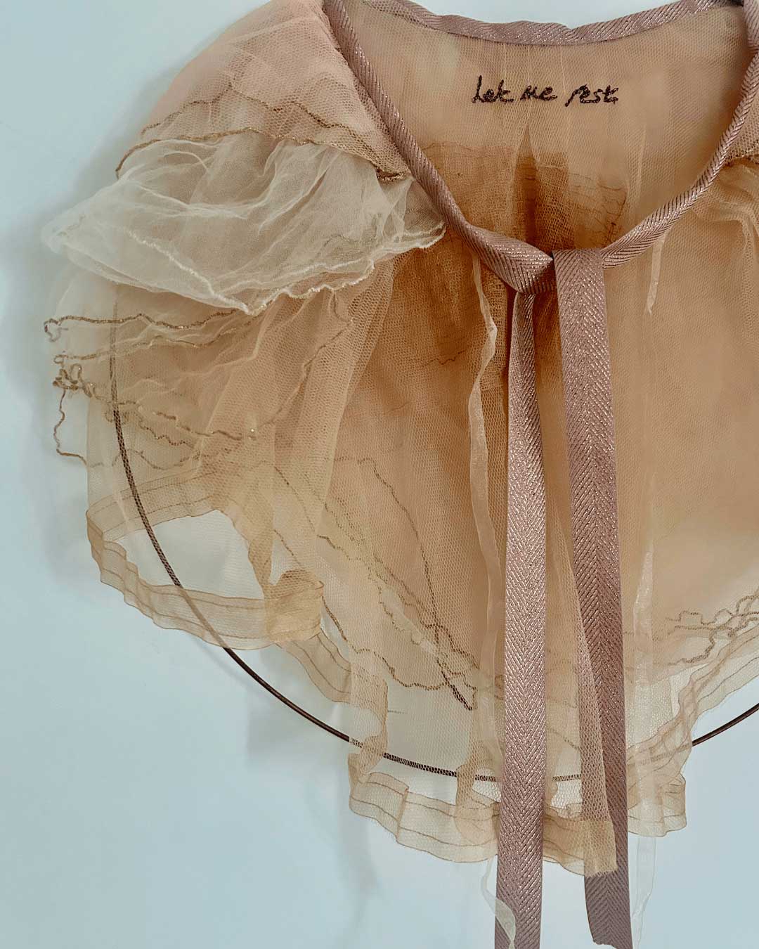 A delicate, sheer, peach-colored ruffled collar with embroidered text 'let me rest' on the neckline and pink ribbon ties, hanging on a thin circular wire.