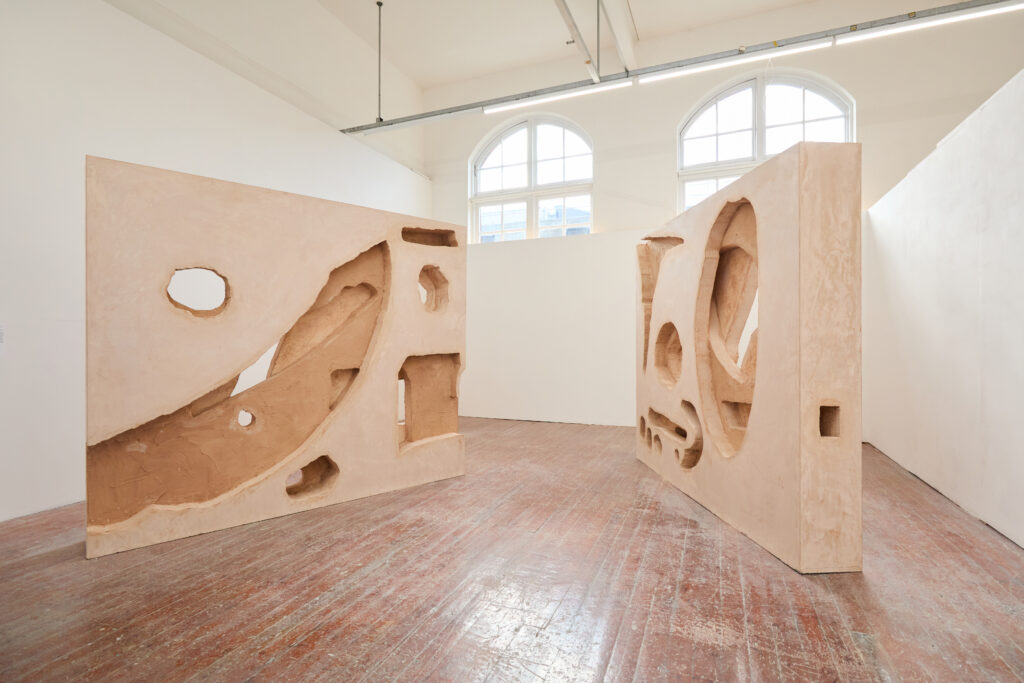 Two large, beige sculptural panels with various geometric cutouts stand upright in a bright art gallery with wooden floors and arched windows, allowing natural light to fill the space.