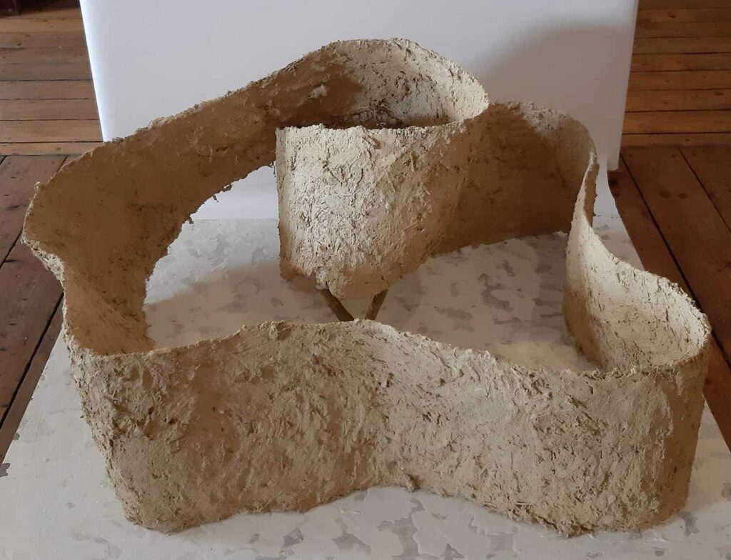 A textured, irregularly-shaped, beige sculpture with uneven surfaces sits on a white platform, set against a wooden floor and a plain white background.